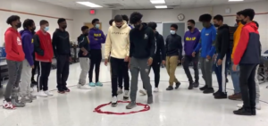 A group of black male high school students are participating in team building experience. They are in a large room and tasked with performing highly coordinated movements in a small space.