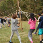 8 multiracial kids in a group pulling ropes to support a person in a center wooden structure